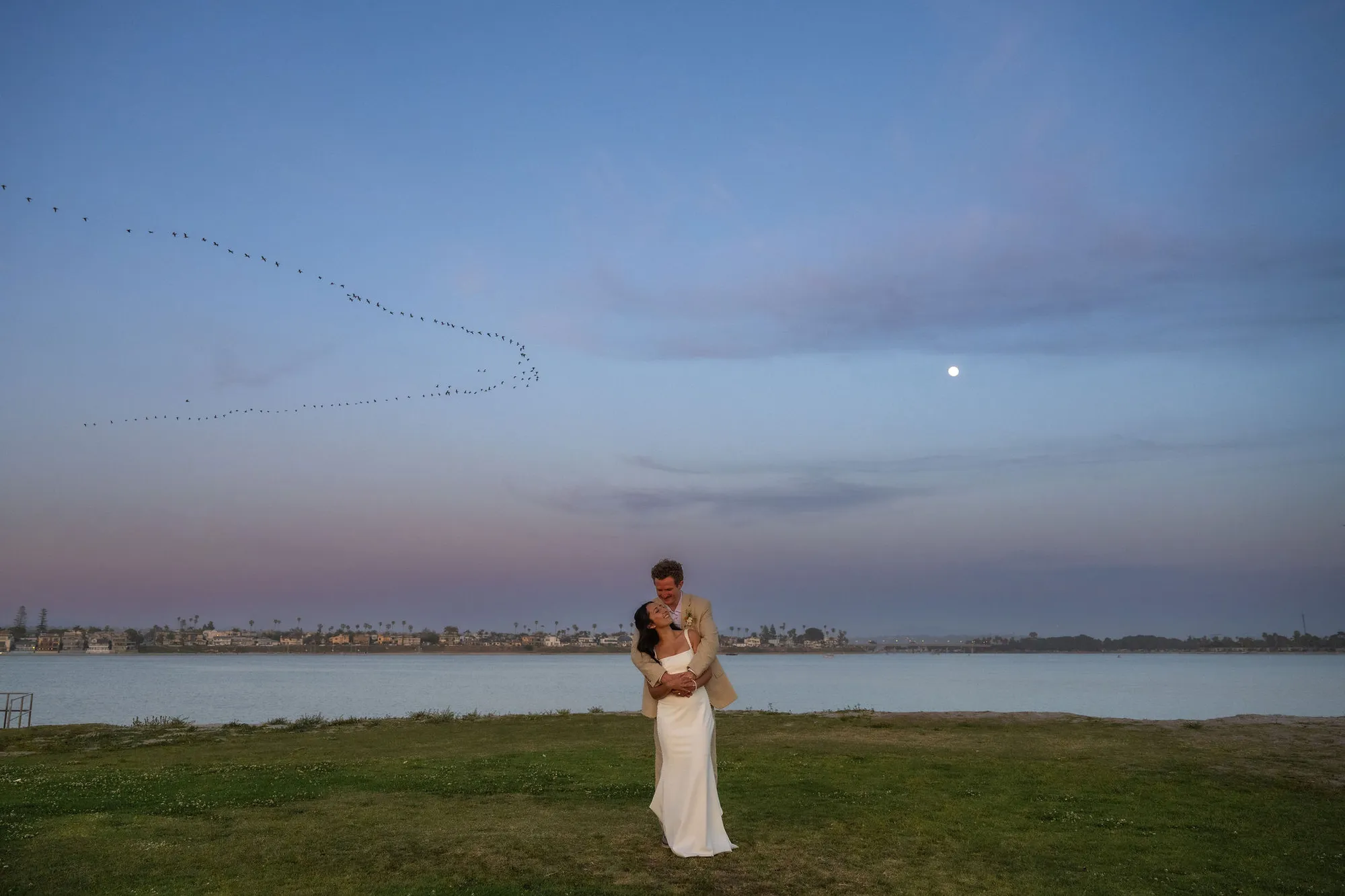 Bride and groom embrace at dusk as birds fly over Mission Bay in San Diego