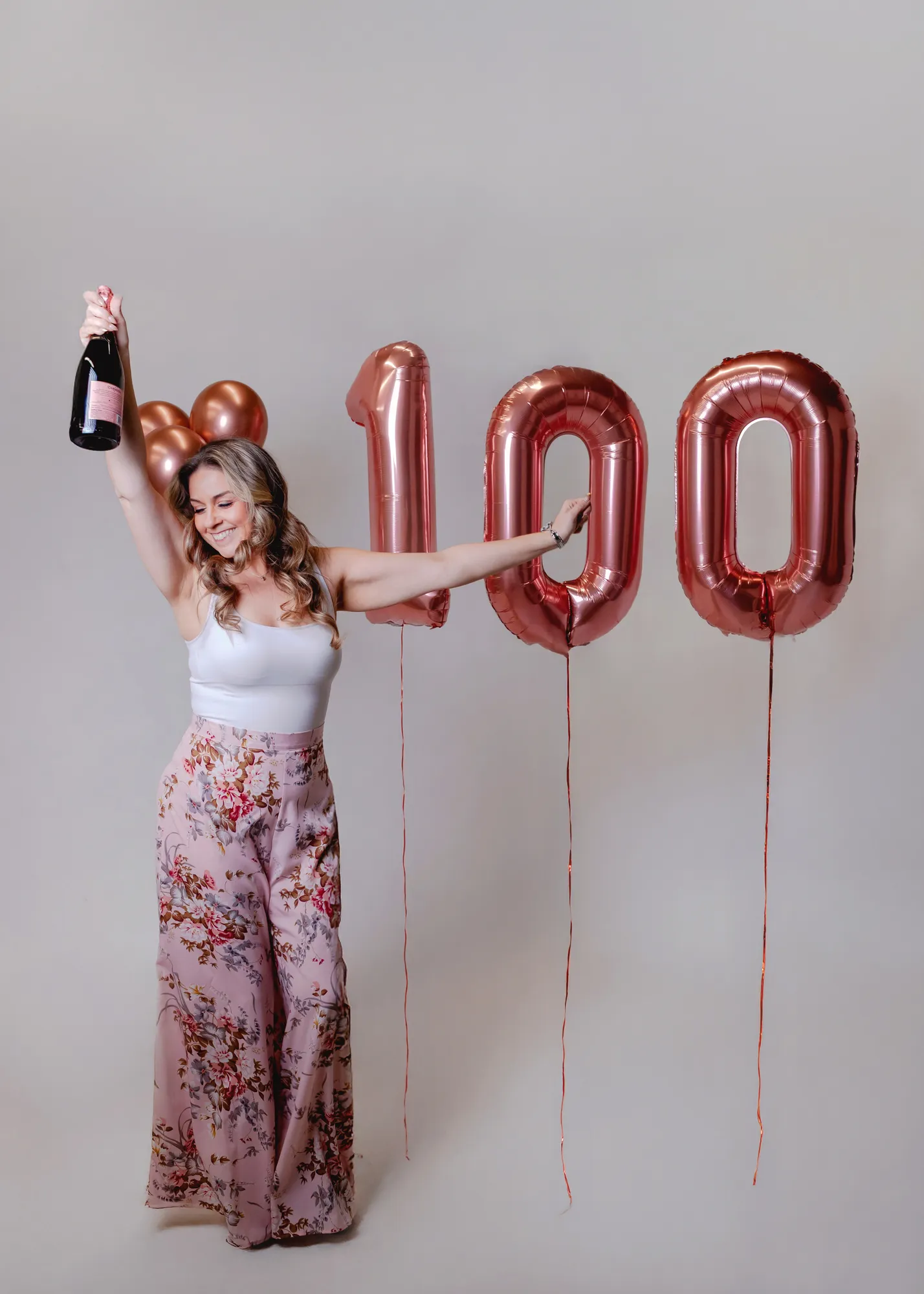 Jen Klaus raising a champagne bottle beside rose-gold 100 balloons, celebrating her 100th wedding